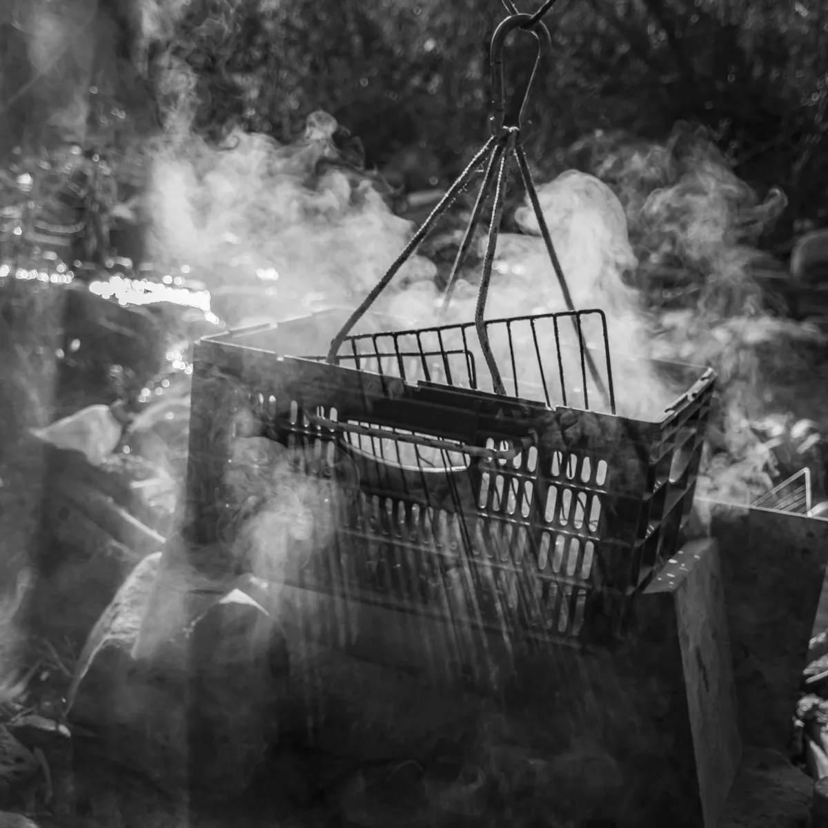 Plastic shopping crate hanging above a smoky outdoor fire in black and white